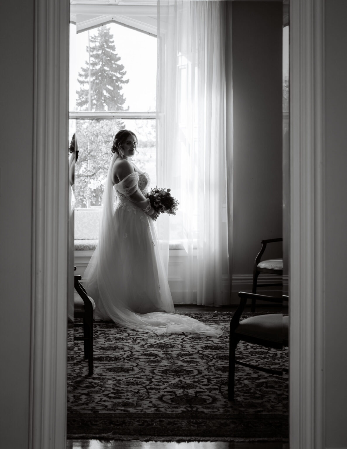 Mariée en robe photographiée en noir et blanc devant une grande fenêtre